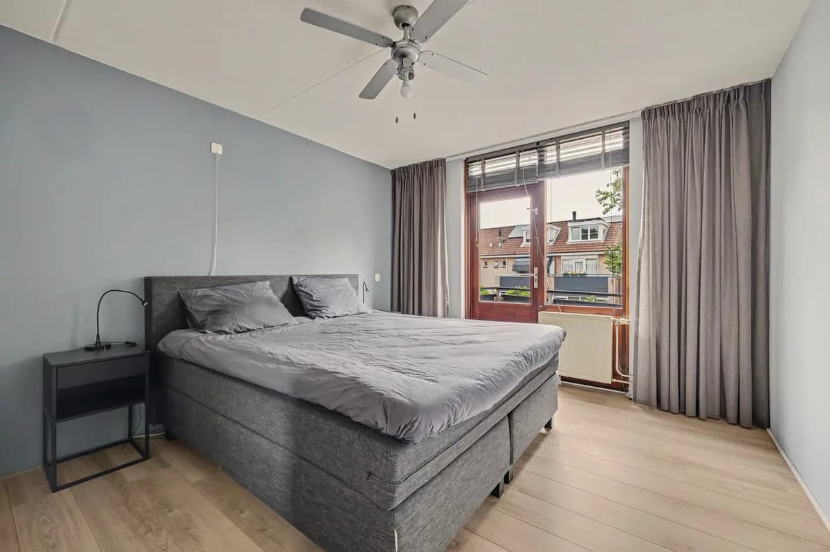 Een gewone slaapkamer met een grijs boxspringbed, een blauwgrijze muur en een raam met gordijnen